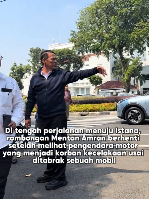 Man in a dark jacket points down the street as he speaks while another person in white stands nearby A city street with trees and parked cars is in the background Indonesian caption overlay reads that a convoy stopped after seeing a motorcyclist who was injured in a crash after being hit by a car