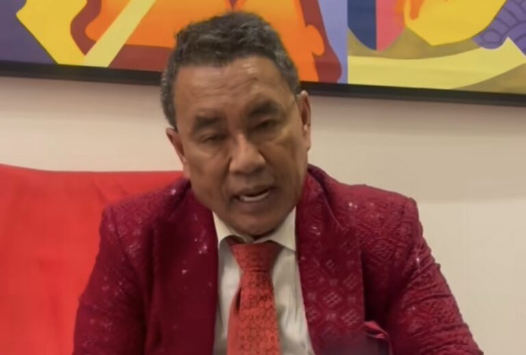 Man in a red patterned suit and red tie speaks, gesturing with his hand while seated on a red sofa.