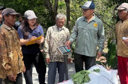 Group of people outdoors gathered around a digital scale weighing green leaves or crops on a tablecloth covered surface