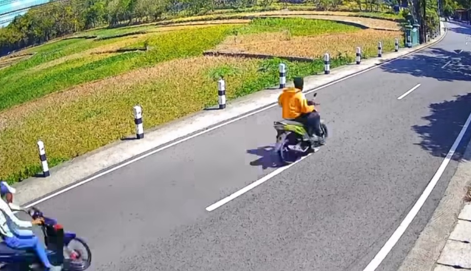 Two motorcyclists ride along a two lane road beside a grassy embankment on a sunny day one rider in an orange shirt is in the center lane while another rider in blue is in the left foreground both wearing helmets