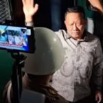 Man in a patterned shirt raises his hand while being recorded by a smartphone on a stand with security personnel nearby