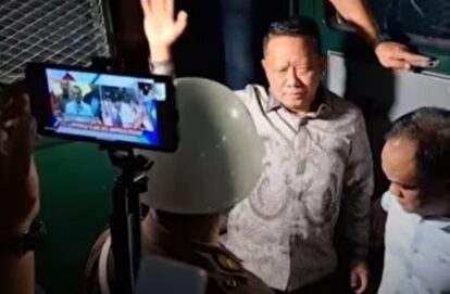 Man in a patterned shirt raises his hand while being recorded by a smartphone on a stand with security personnel nearby