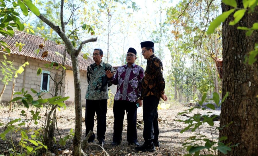 Three men in batik shirts and peci hats stand in a sunlit tree filled yard beside a small brick building talking