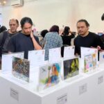 Visitors examine colorful artworks displayed on white pedestals at an art exhibition in a bright gallery setting with a busy crowd