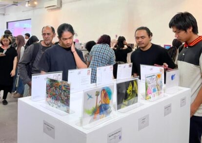 Visitors examine colorful artworks displayed on white pedestals at an art exhibition in a bright gallery setting with a busy crowd