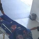 Two emergency responders push a patient on a wheeled stretcher through a clinic the patient covered with a blue floral blanket in a hallway