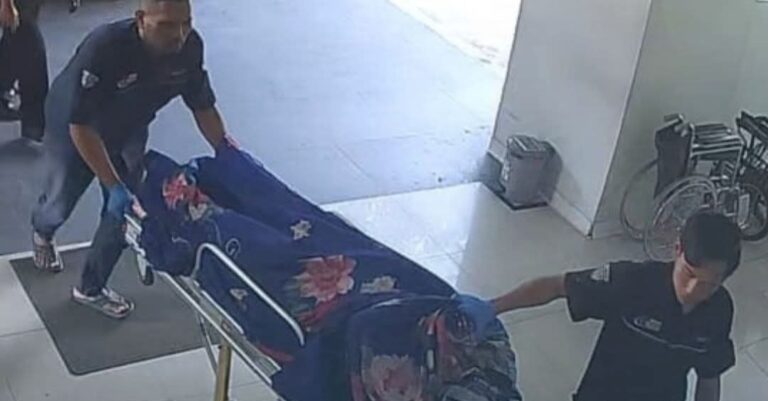 Two emergency responders push a patient on a wheeled stretcher through a clinic, the patient covered with a blue floral blanket in a hallway.