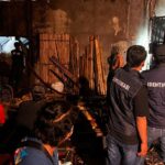 Investigators in dark vests labeled identifikasi examine a dim cluttered workshop or construction site with debris and tools scattered around