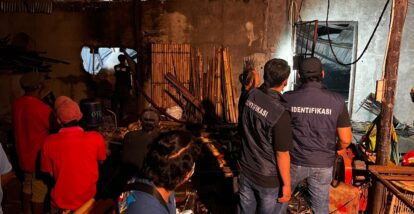 Investigators in dark vests labeled identifikasi examine a dim cluttered workshop or construction site with debris and tools scattered around