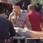 Police and medical responders carry a person on a stretcher through a crowded outdoor area