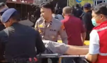 Police and medical responders carry a person on a stretcher through a crowded outdoor area
