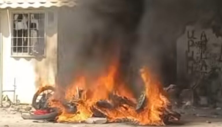 A pile of burning tires and debris in front of a beige building, with thick black smoke rising from the flames.