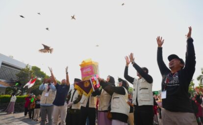 People outdoors in a crowd raise their hands in celebration as birds fly overhead a central figure holds a bright yellow bag or banner with pink text