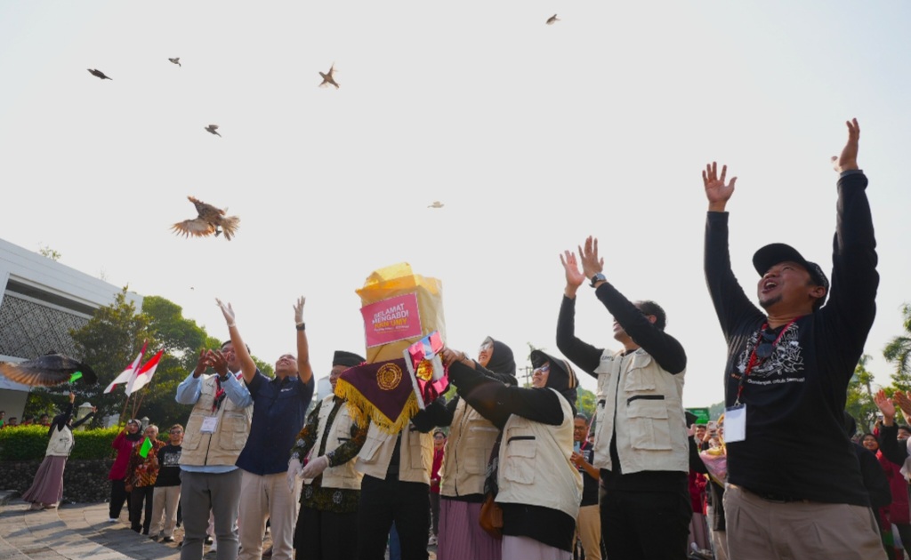 People outdoors in a crowd raise their hands in celebration as birds fly overhead a central figure holds a bright yellow bag or banner with pink text