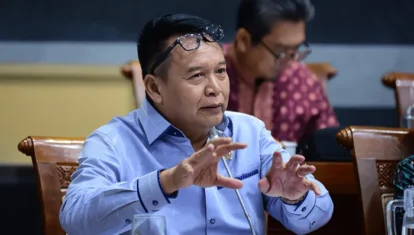 Man in a light blue shirt gestures with hands while speaking at a meeting, glasses perched on his head, others seated in the background