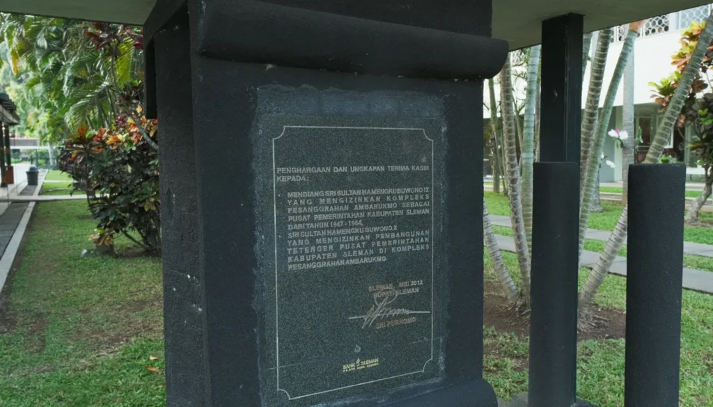 Tetenger12048x1170 catrawarta Black stone monument with an engraved plaque in indonesian set in a landscaped garden with palm trees nearby