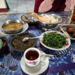 A table spread with rice greens curry and a cup of tea seen from above as a person sits at the table in the background