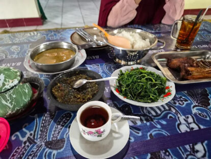 A table spread with rice, greens, curry, and a cup of tea, seen from above as a person sits at the table in the background.