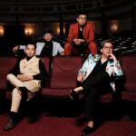 Four men in stylish outfits pose in a dim theater with red seats behind them