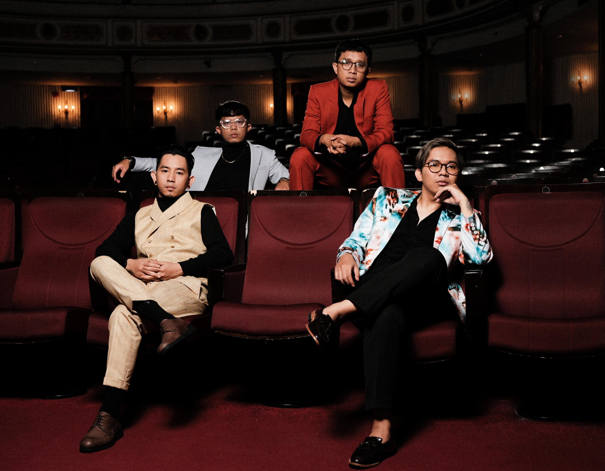 Four men in stylish outfits pose in a dim theater with red seats behind them