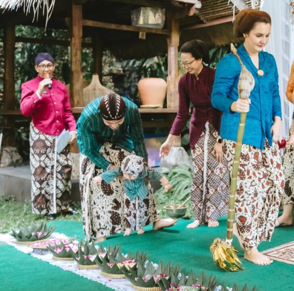 Group of women in batik sarongs and colorful blouses participate in a traditional outdoor ceremony guiding a child near decorative leaf offerings on a green carpet