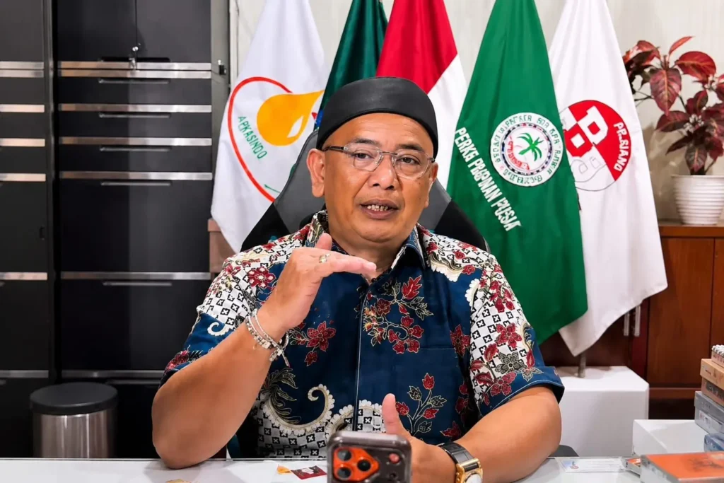 Man in a batik shirt and black cap speaks at a desk with multiple flags in the background