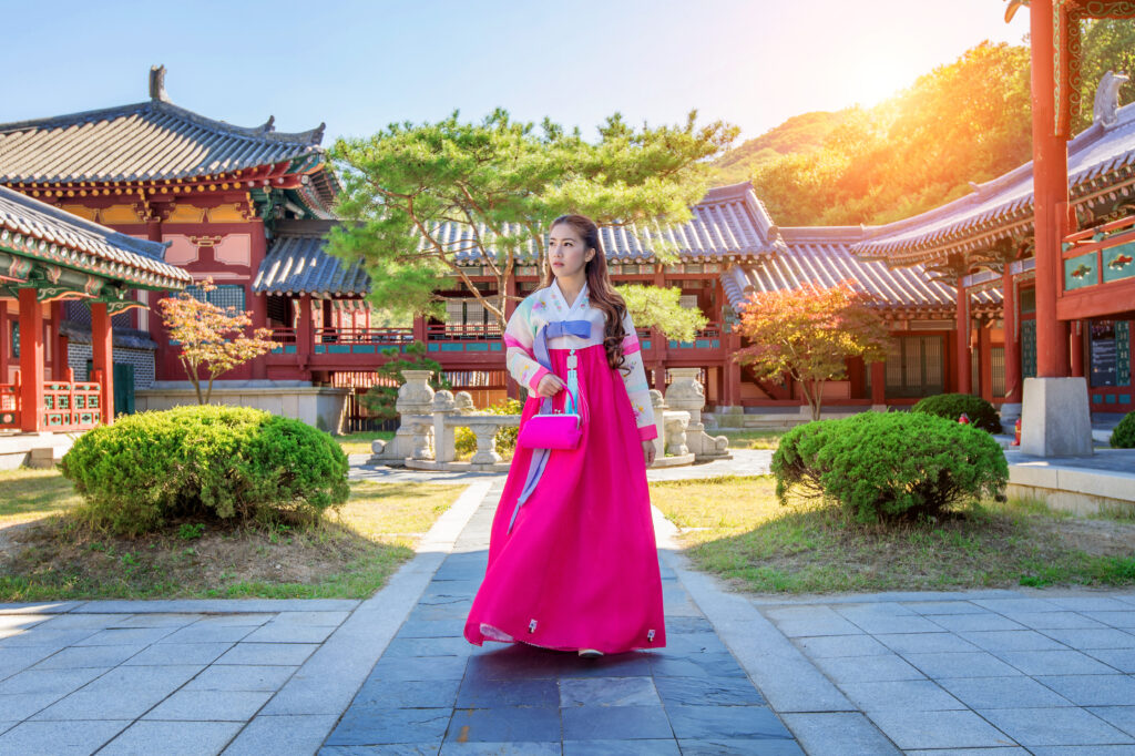 Woman in a pink and white hanbok stands in a traditional korean palace courtyard with red wooden buildings and tiled roofs