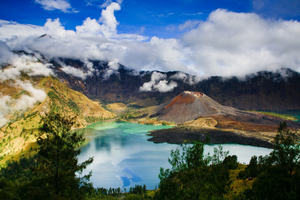 Turquoise crater lake surrounded by a volcanic cone and green hills under a cloudy sky